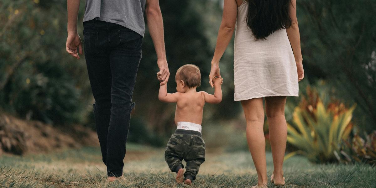 man and woman walk away from camera holding hands of baby walking in between them